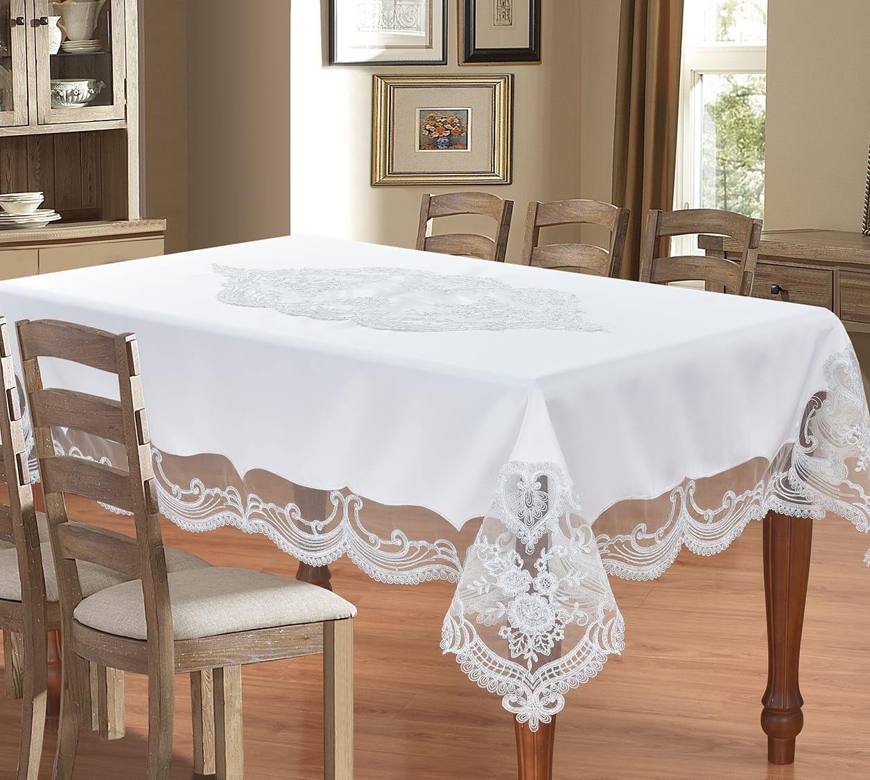 A white rectangular tablecloth with an embroidered floral motif in the center and scalloped lace trim along the edges, shown draped over a wooden dining table in a furnished dining room.