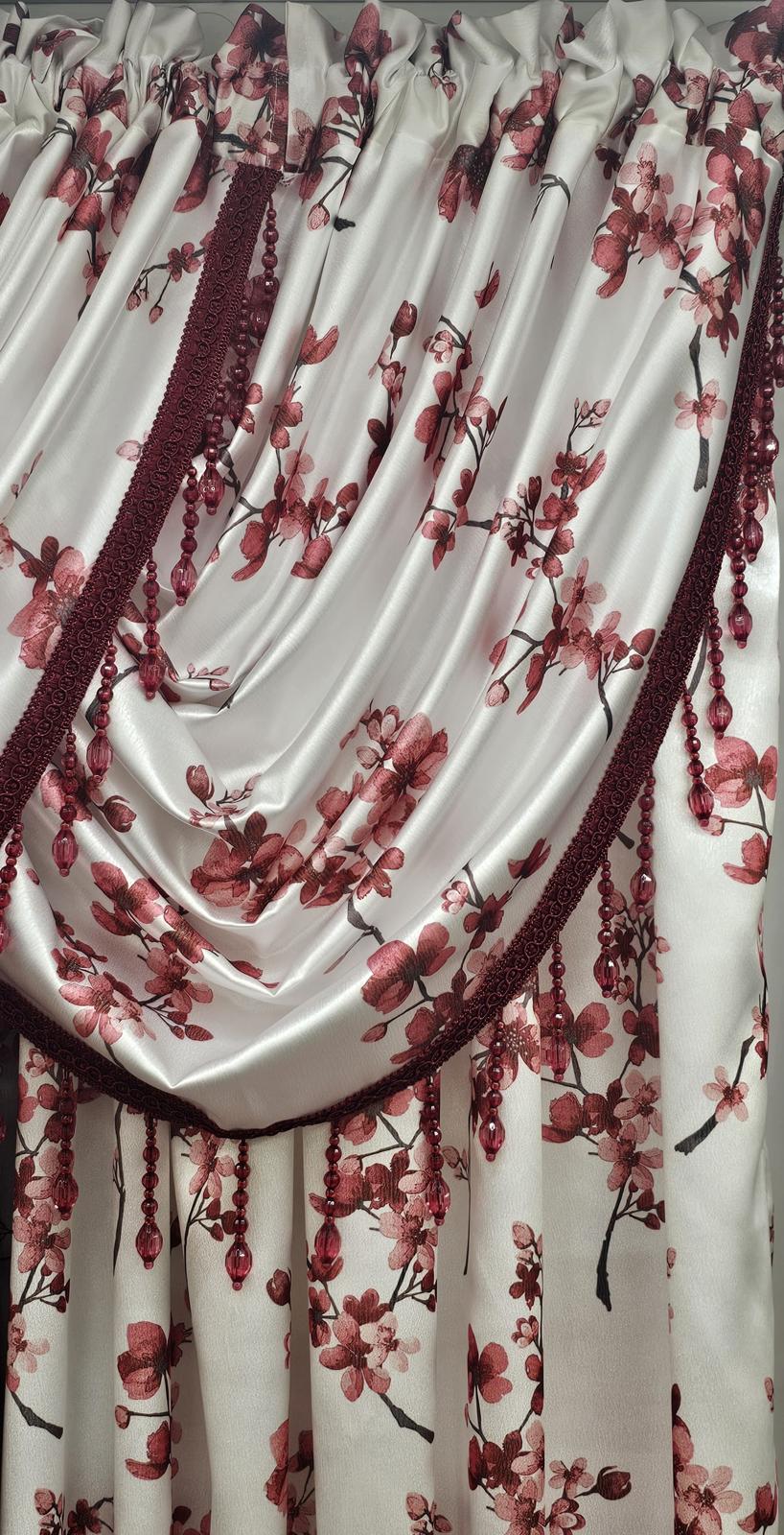 A close-up of a draped curtain panel with a cream satin-like fabric, decorated with a red and pink cherry blossom floral pattern and dark burgundy lace trim.