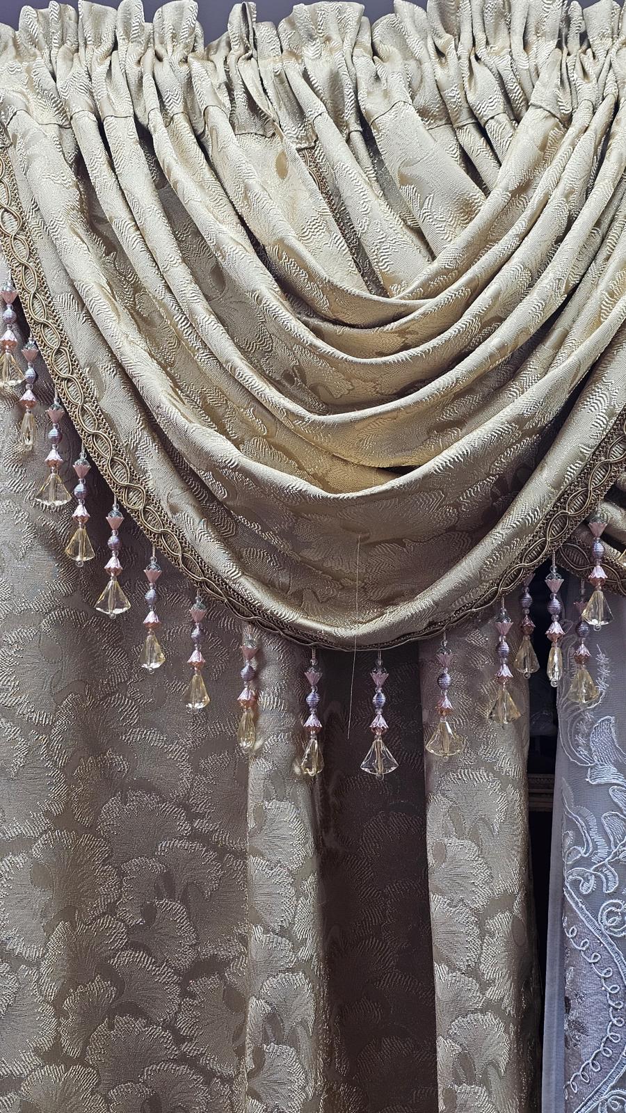 A close-up of an ornate beige and gold damask curtain valance with cascading crystal bead tassels hanging from the scalloped edge.