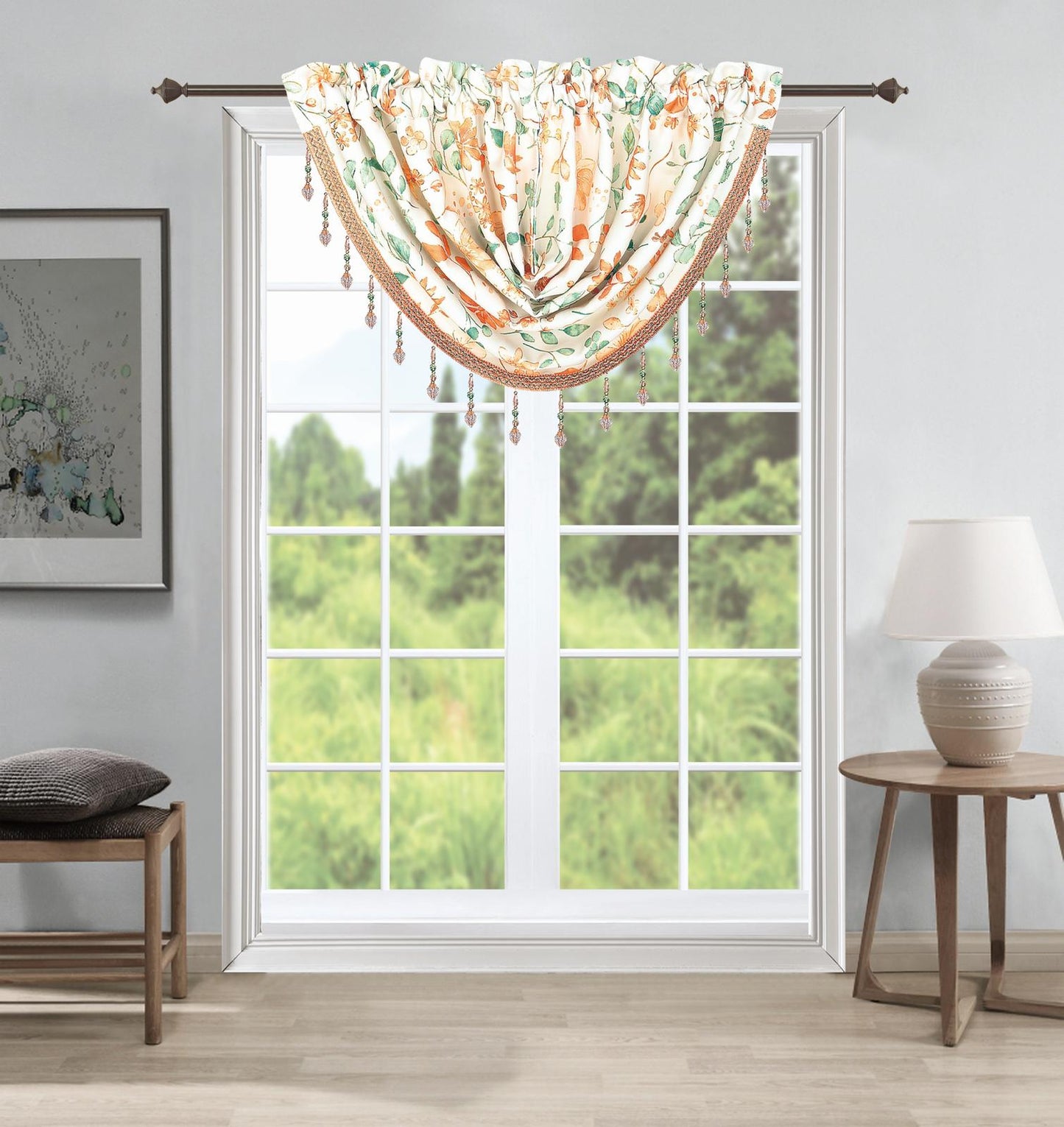 A floral valance curtain with tassel fringe hanging from a dark metal rod over a white multi-pane window. The room setting includes a light gray wall, a wooden stool with a cushion on the left, and a wooden side table with a lamp on the right, looking out onto a green grassy field.