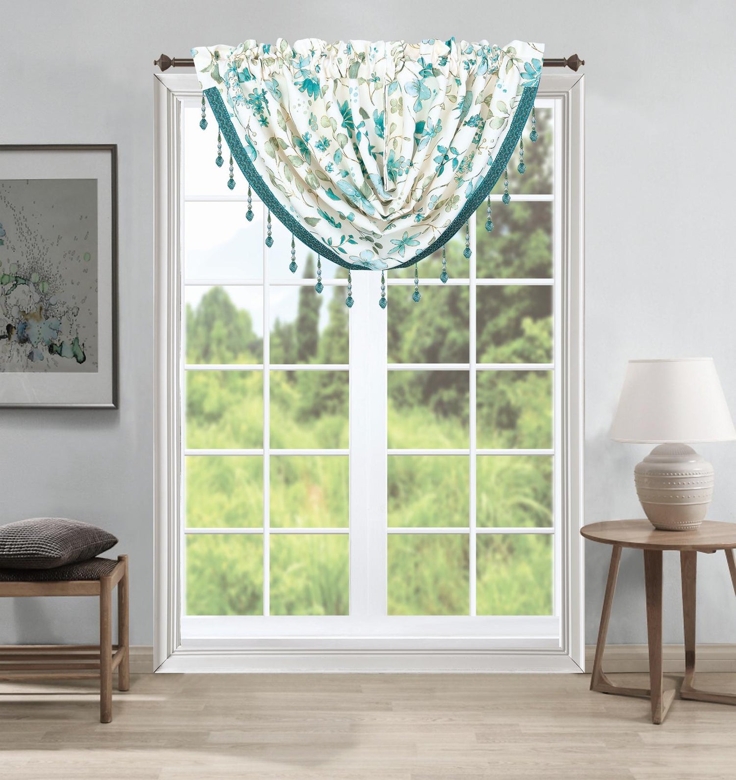 A decorative window valance with teal and cream floral patterns and hanging bead tassels, shown hanging on a window in a styled room with a side table, lamp, and bench.