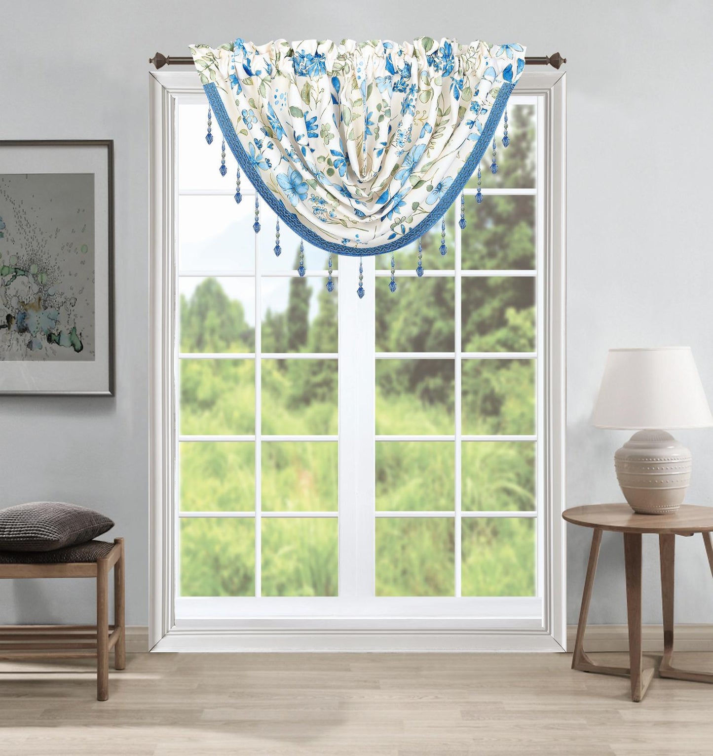 A blue and white floral valance with hanging tassels is shown hanging on a window in a styled room with a side table, lamp, and bench.