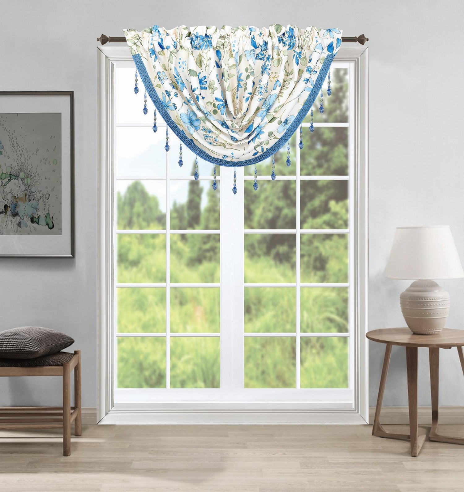 A blue and white floral valance with hanging tassels is shown hanging on a window in a styled room with a side table, lamp, and bench.