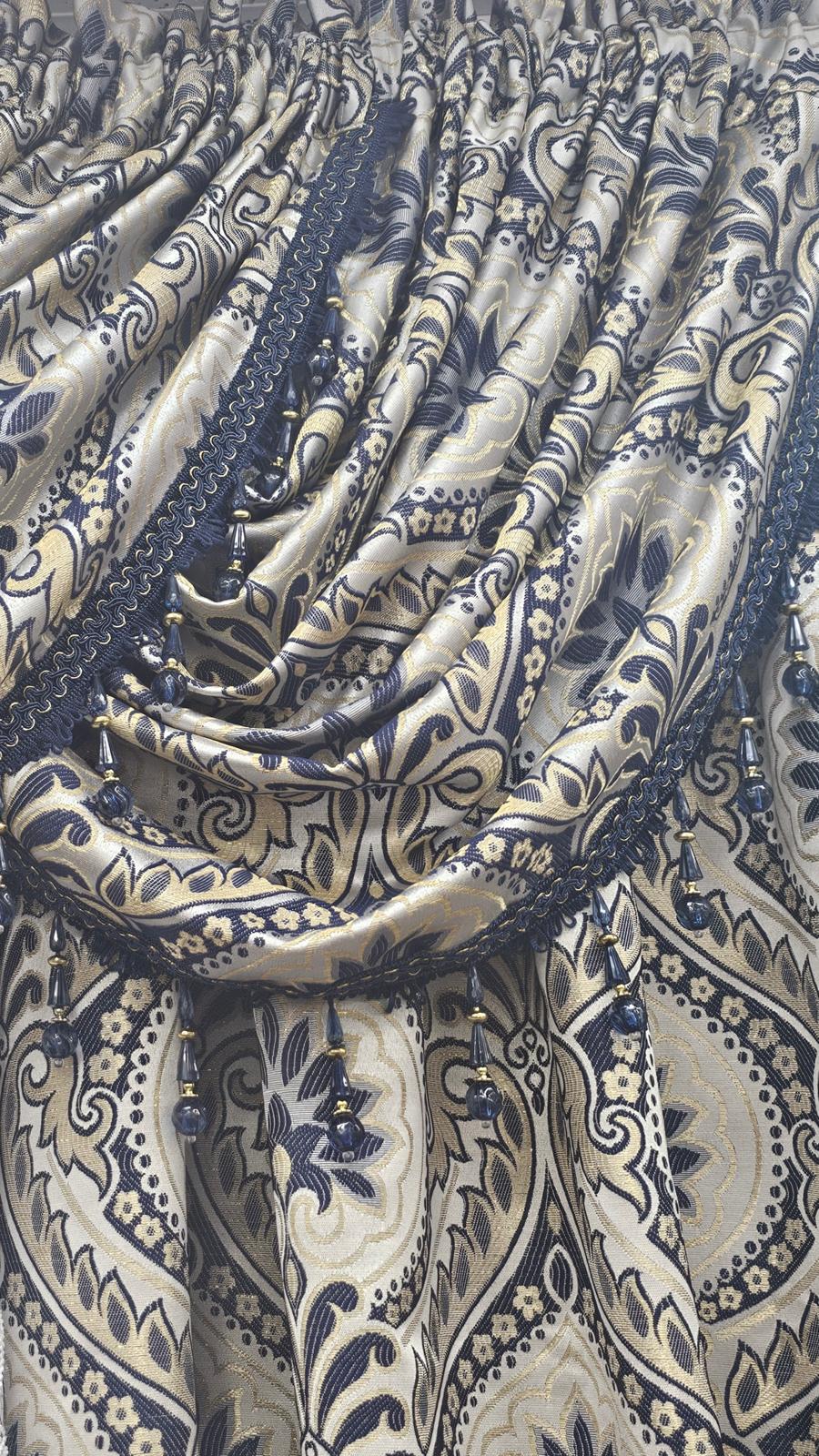 Close-up of a gathered curtain panel with a beige and navy blue paisley pattern, featuring decorative dark blue lace trim and hanging bead tassels.