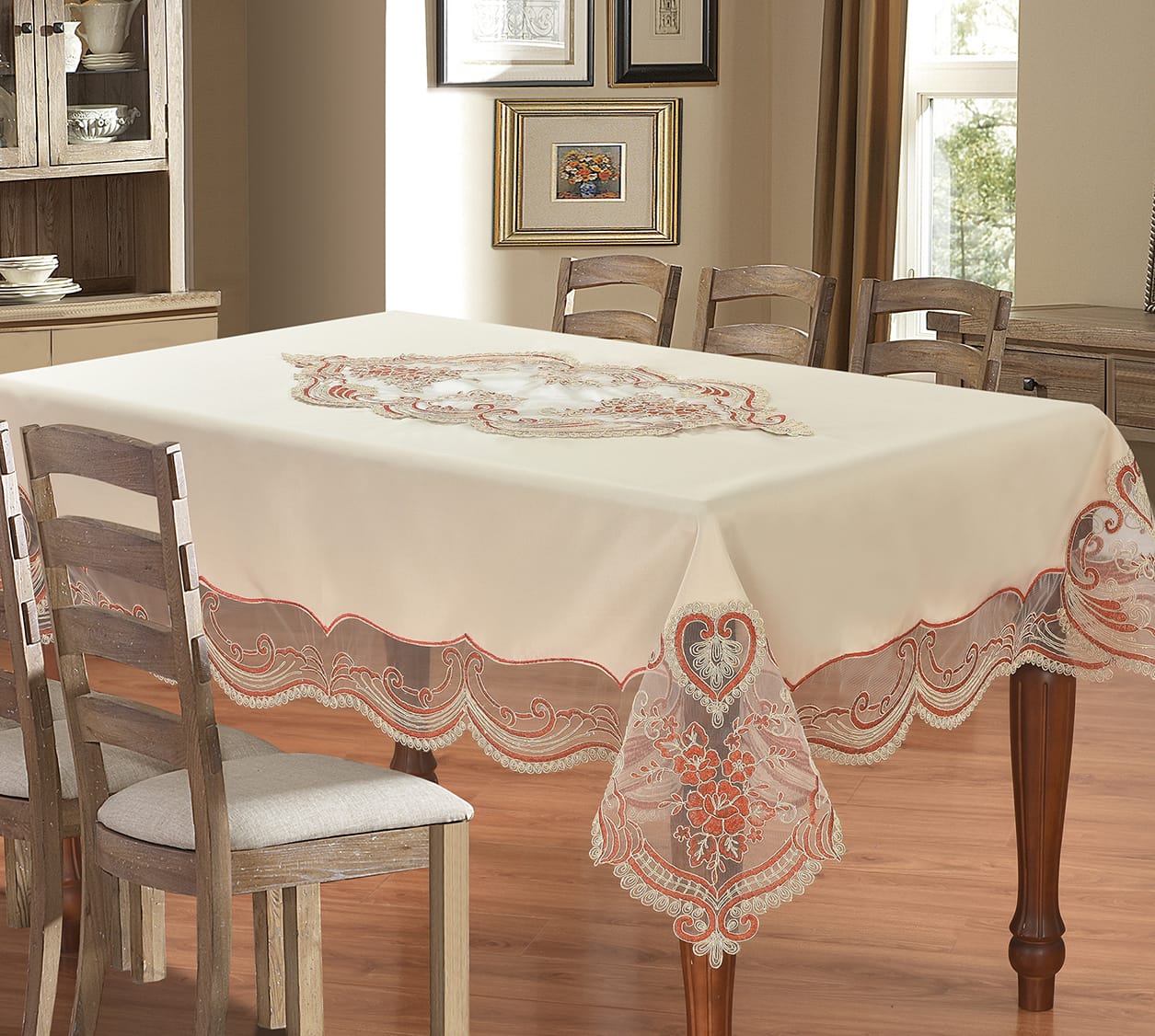 An off-white tablecloth with ornate embroidered floral and scroll patterns and scalloped lace-like borders, draped over a wooden dining table in a warm, traditional dining room setting.