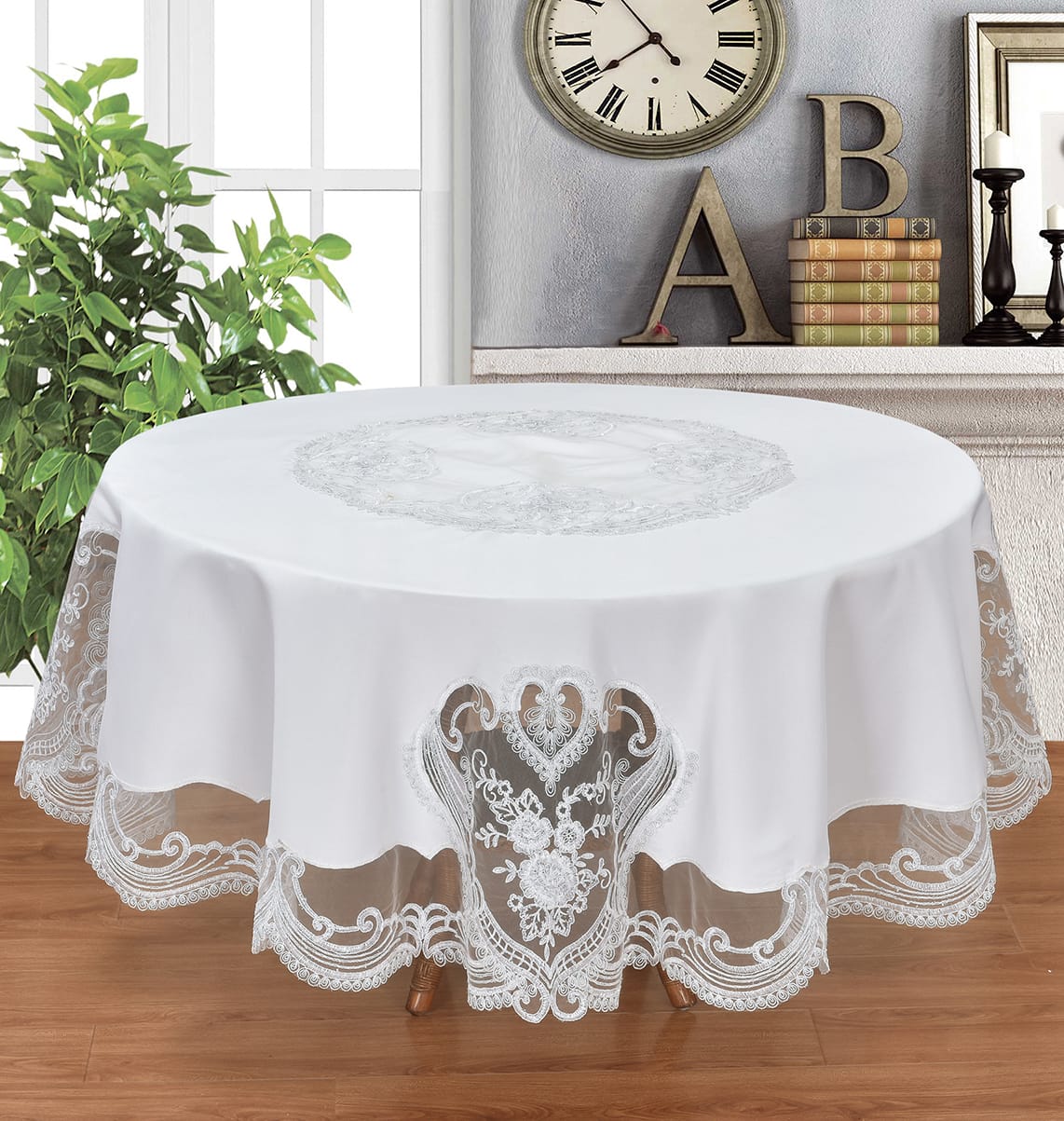 A round white tablecloth with an embroidered floral motif in the center and a scalloped lace hem, draped over a table in a decorated room with a plant, wall clock, and decorative letters.