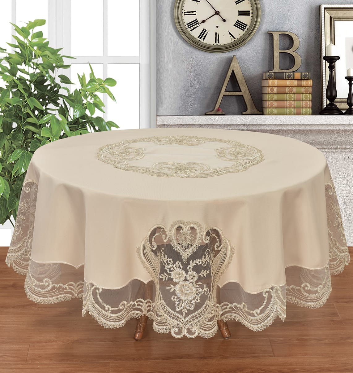 A round beige tablecloth with intricate floral embroidery and lace trim, draped over a table in a decorated room with a clock, books, and a plant.