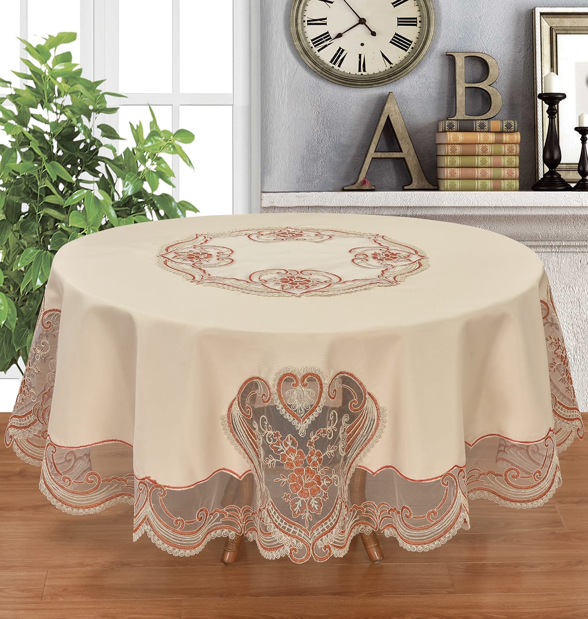 Round tablecloth in warm cream with ornate embroidered floral and scroll motifs in rust orange and taupe, draped over a table in a styled room with wood flooring, a plant, wall decor, and a window.